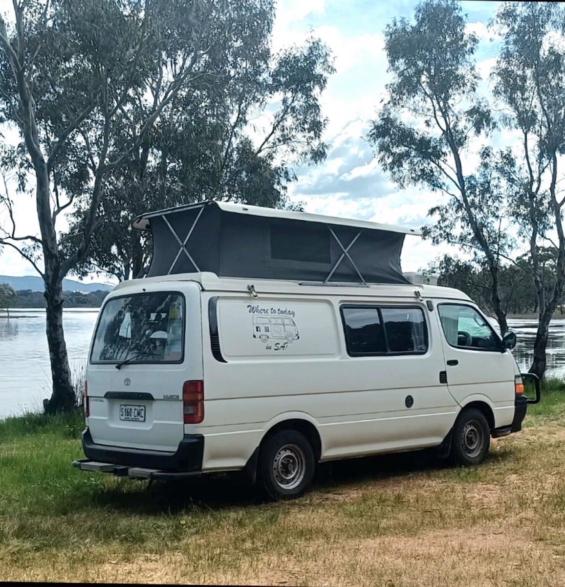 Trevor the campervan with its pop-top roof raised, parked on grass between gum trees at the edge of a lake.